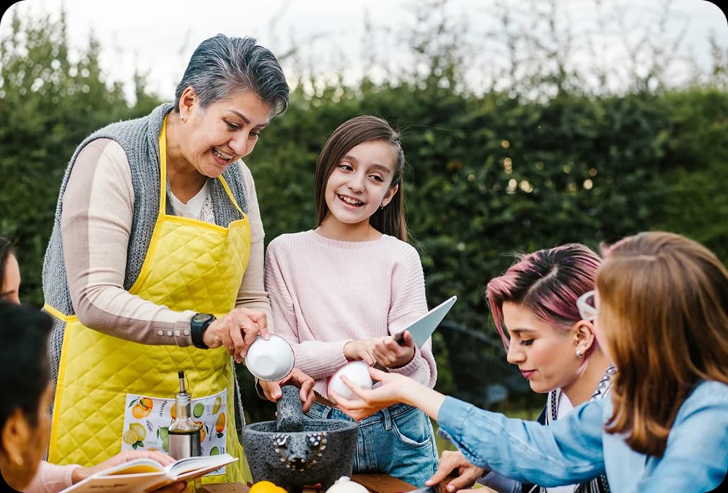 Un adulto enseña a los niños a preparar ingredientes con un mortero durante una actividad de aprendizaje cultural práctica al aire libre.