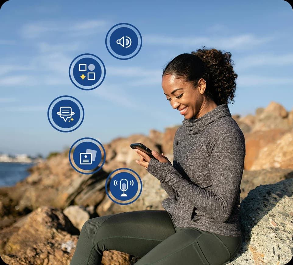 Una mujer sentada al aire libre sobre rocas cerca del agua, sonriendo y mirando su teléfono, con iconos de funciones de Rosetta Stone Sapphire superpuestos a su alrededor.